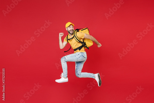 Full length delivery employee man guy male 20s in yellow cap t-shirt uniform thermal food bag backpack work courier service during quarantine covid-19 virus jumping isolated on red background studio.