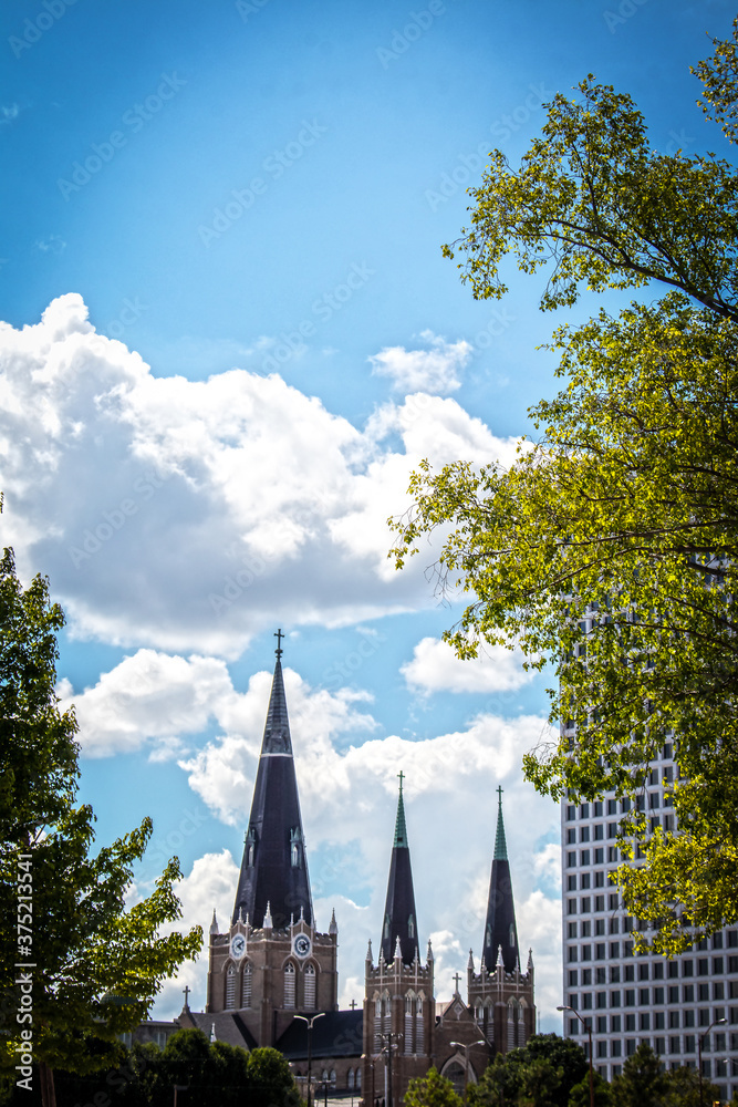 Spires of Tulsa with modern and older buildings framed by trees under a beautiful blue skies with fluffy clouds