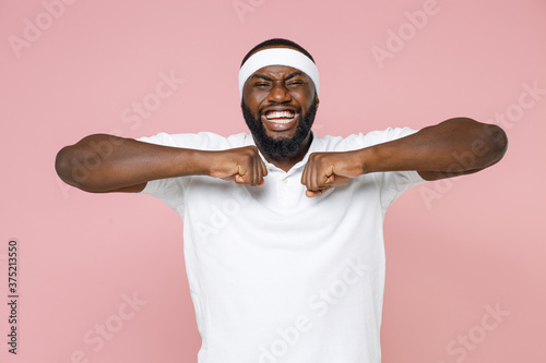 Cheerful young bearded african american fitness sports man 20s in white headband t-shirt doing stretching exercising spending time in gym isolated on pastel pink color wall background studio portrait.