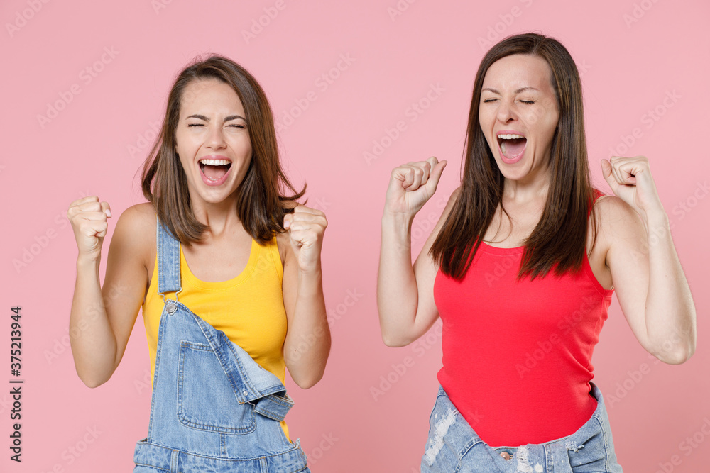 Two happy joyful young women friends 20s in casual denim clothes posing standing clenching fists doing winner gesture keeping eyes closed isolated on pastel pink colour background, studio portrait. Two happy joyful young women friends 20s in casual denim clothes posing standing clenching fists doing winner gesture keeping eyes closed isolated on pastel pink colour background, studio portrait.
