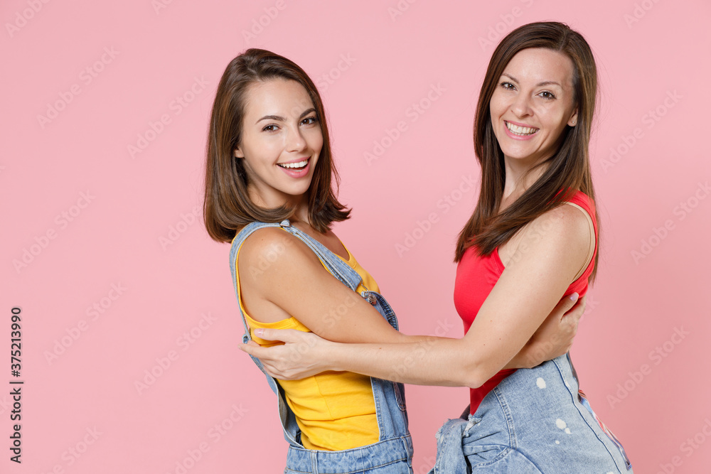 Side view of two cheerful smiling beautiful attractive young women friends 20s wearing casual denim clothes hugging looking camera isolated on pastel pink colour background, studio portrait. Side view of two cheerful smiling beautiful attractive young women friends 20s wearing casual denim clothes hugging looking camera isolated on pastel pink colour background, studio portrait.