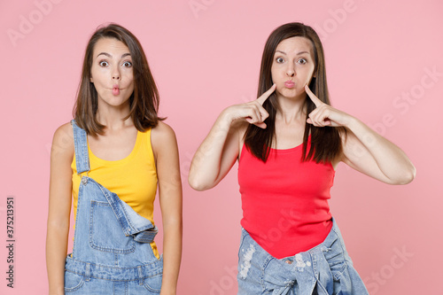Two funny shocked young women friends 20s in denim clothes pointing index fingers on blowing puffing cheeks with funny face mouth inflated with air isolated on pink colour background, studio portrait.