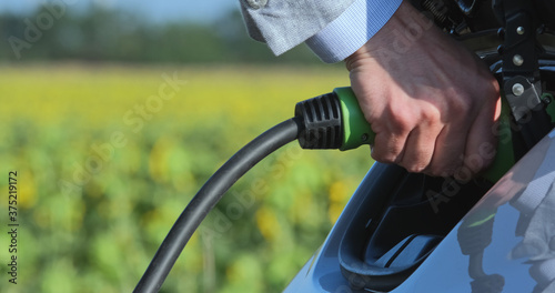 Close-up of a man inserting a charger into his electric car against the backdrop of a green sunflower field.