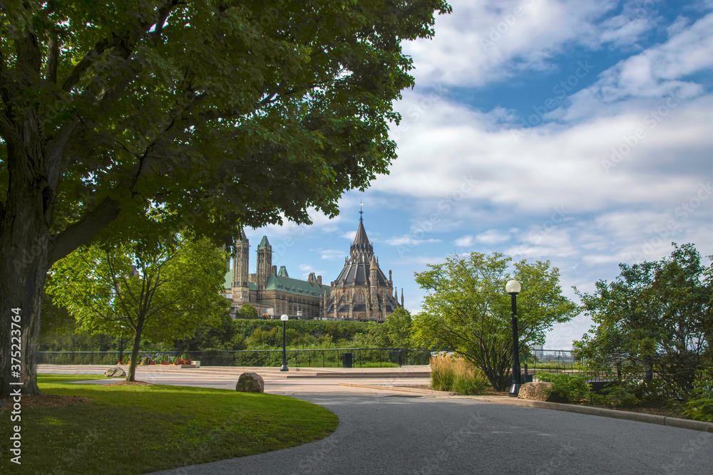 View of Canadian Parliament from Major's Hill Park under blue cloudy skies with long sweeping walkways, lawns, trees and lampposts in foreground nobody