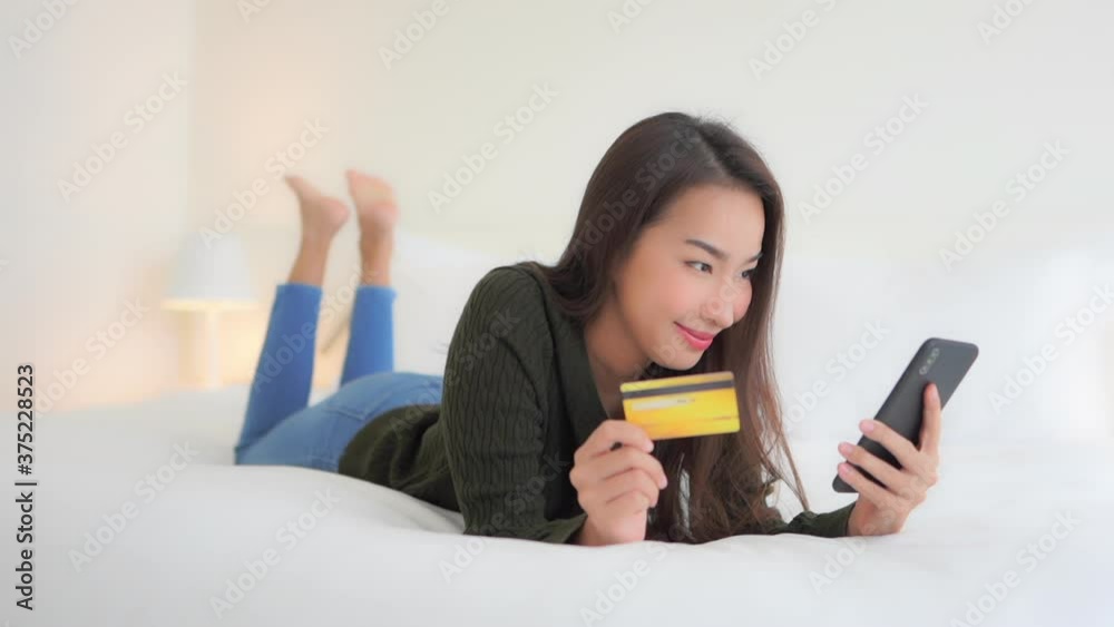 Beautiful Thai woman lying on king-size luxury bed and type in the credit card number to her mobile phone to pay online Beautiful Thai woman lying on king-size luxury bed and type in the credit card number to her mobile phone to pay online