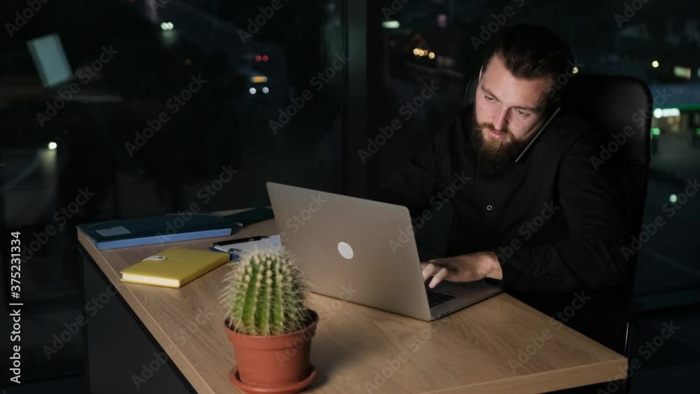 Bearded businessman talking at the phone with his wife while is working in a modern office at midnight. Bearded businessman talking at the phone with his wife while is working in a modern office at midnight.