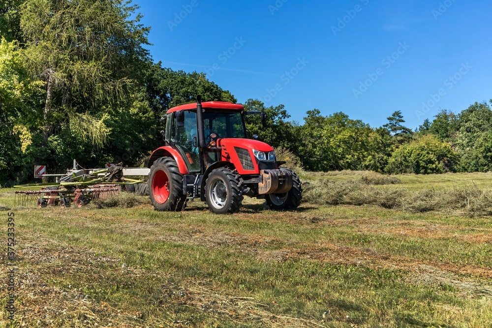 Red tractor working in a field with a two-rotor tedder. Drying hay in the meadow. Work on an agricultural farm. Red tractor working in a field with a two-rotor tedder. Drying hay in the meadow. Work on an agricultural farm.