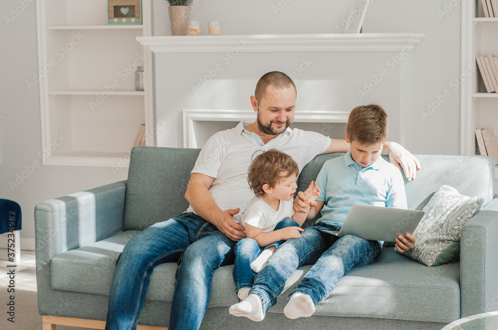 Happy young father sit on couch using laptop relax with preschooler son holding smartphone have fun together, smiling dad and little boy child enjoy weekend at home rest on sofa busy with gadgets