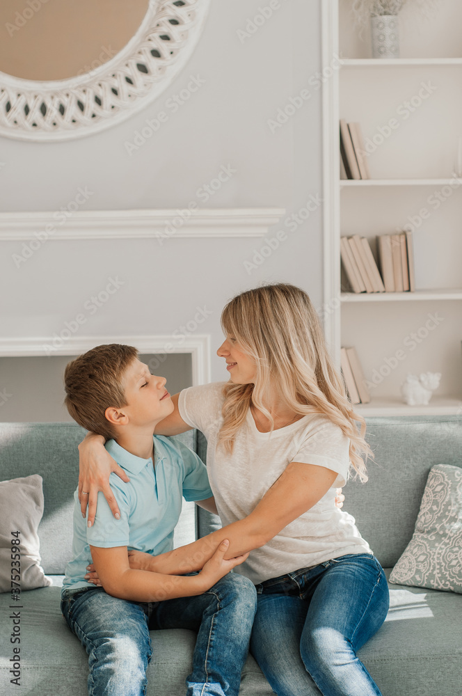Mom and kid are laughing together in cozy home. They are holding each other with joy while relaxing on sofa. Parent is playfully touching boy feet and he is enjoying it