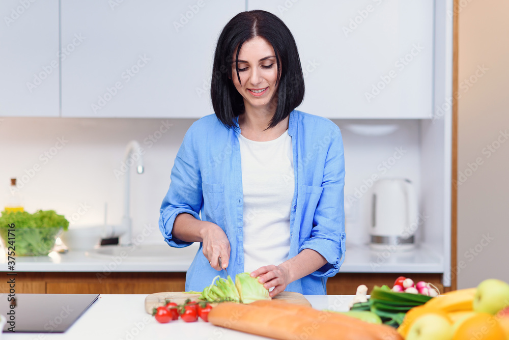 Young woman cuts the cabbage with a knife on a wooden board and preparing salad from vegetables at cozy home kitchen.