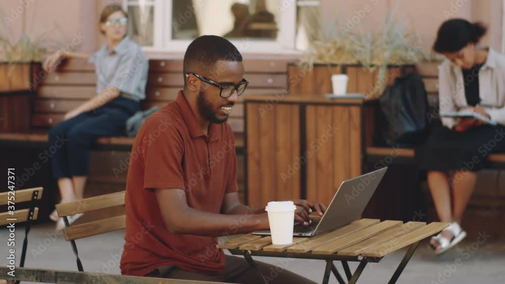 Young Afro-American man sitting at table in outdoor street cafe with disposable coffee cup, smiling and working on laptop