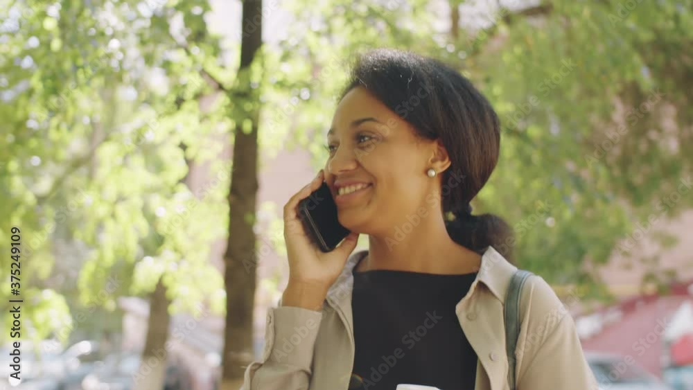 Chest-up shot of beautiful African American woman holding coffee to go cup, walking through park on summer day, smiling and chatting on mobile phone Chest-up shot of beautiful African American woman holding coffee to go cup, walking through park on summer day, smiling and chatting on mobile phone