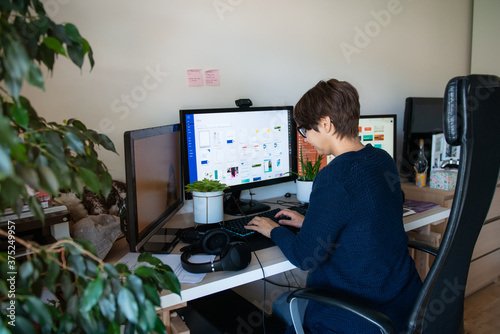 Back view woman in glasses typing on pc with headsets, few monitors on her work desk at home working space. Remote work concept. Freelancer workspace. Selective focus. Copy space