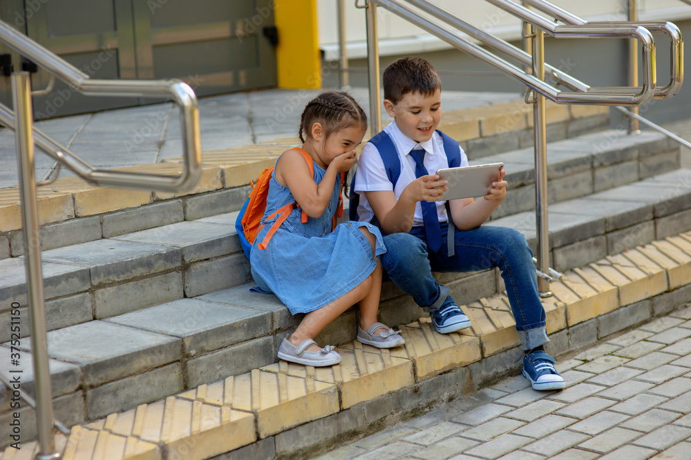 Two schoolchildren a girl with an orange backpack and a boy , laugh, sit on the stairs outside and look into a tablet Two schoolchildren a girl with an orange backpack and a boy , laugh, sit on the stairs outside and look into a tablet
