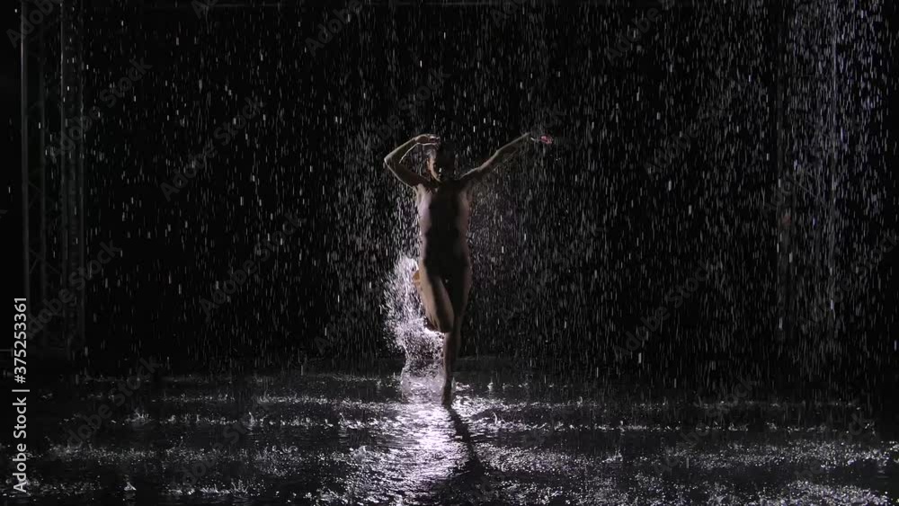 Impulsive female artist dancing in modern style of contemporary choreography against black background with backlight. Young expressive actress performing energetic dance moves in the rain.