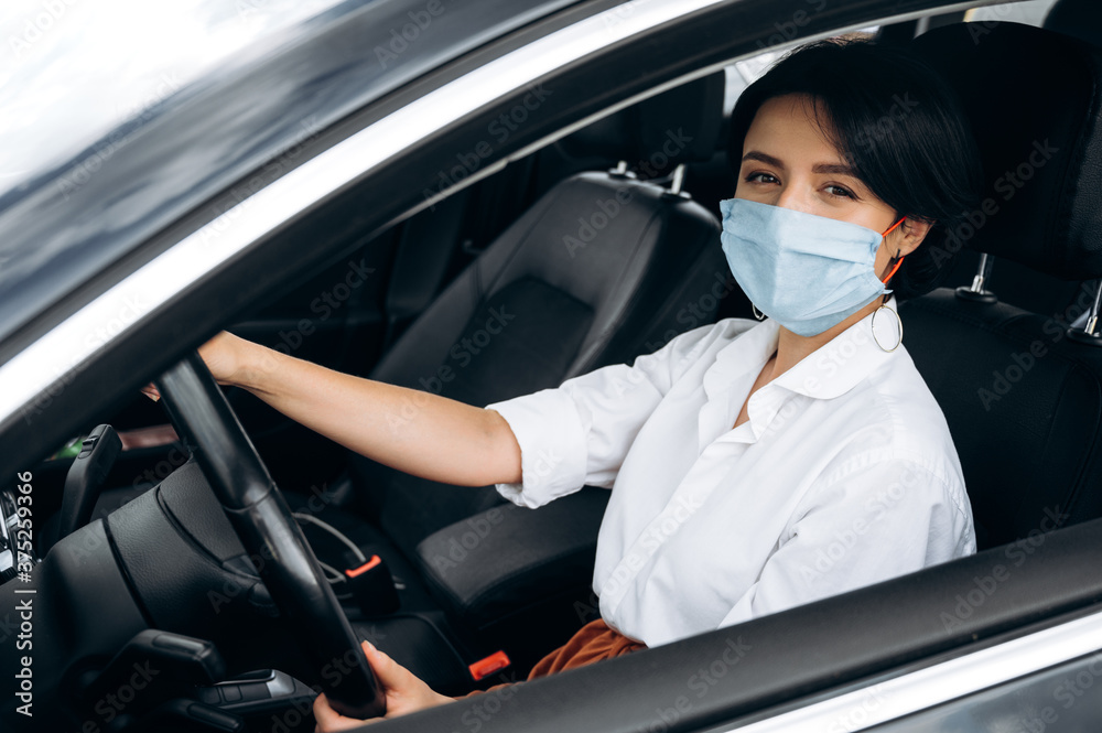 Stay safety. A girl in a protective medical mask sits behind the wheel of his car and looks at the camera Stay safety. A girl in a protective medical mask sits behind the wheel of his car and looks at the camera