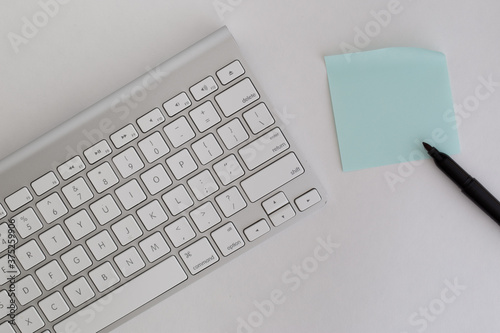 Wireless computer keyboard on white background with light blue post it note next to it with space for copy text and a black pen