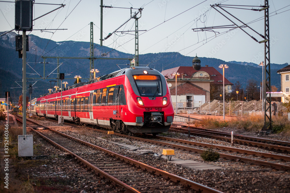 Red commuter train is entering a station of Garmisch Partenkirchen in evening hours, with fantastic background view over the mountains and snowy hills.