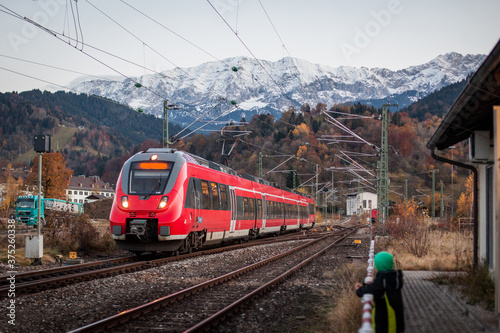 Red commuter train is entering a station of Garmisch Partenkirchen in evening hours, with fantastic background view over the mountains and snowy hills.