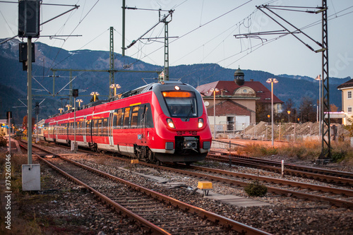 Red commuter train is entering a station of Garmisch Partenkirchen in evening hours, with fantastic background view over the mountains and snowy hills.