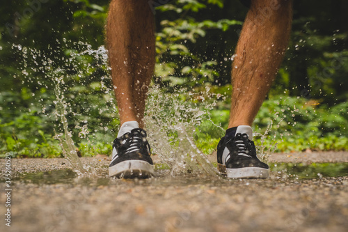 Male legs jumping into a small pond on tarmac, creating a big splash of clear water that is coming out from below sport shoes which the man is wearing. Dark sport shoes landed in water.
