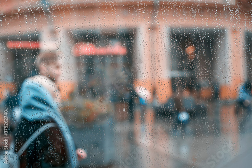 Autumn rainy weather, walking happy young couple together. View through a wet window with raindrops. Concept of modern city, love, lifestyle. Abstract blurred background