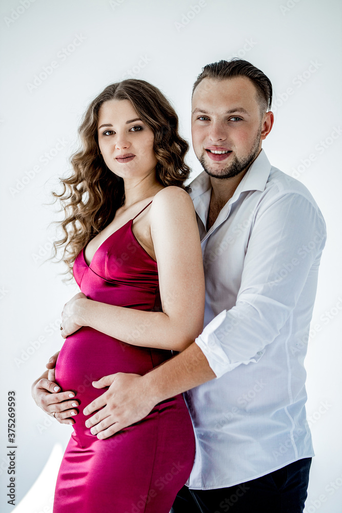 A beautiful curly pregnant girl in an evening dress stands in an embrace with her husband on a white background. She has a crimson evening dress and he is wearing festive white shirt. Family Concept A beautiful curly pregnant girl in an evening dress stands in an embrace with her husband on a white background. She has a crimson evening dress and he is wearing festive white shirt. Family Concept