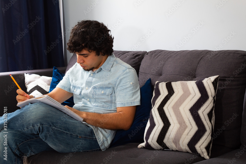 Young hispanic man with wavy hair studying from home writing in a notebook with a pencil, sitting on a sofa