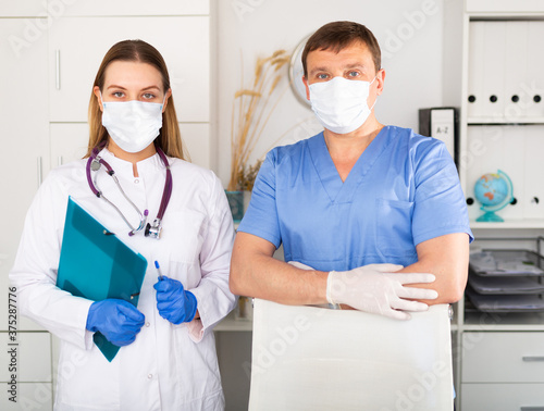 Portrait of two professional qualified health workers wearing disposable surgical masks and latex gloves in modern medical office