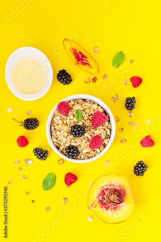 Crunchy granola, berries and mint in ceramic bowl and raspberries, blackberries, peach and honey on yellow background. Healthy breakfast concept.