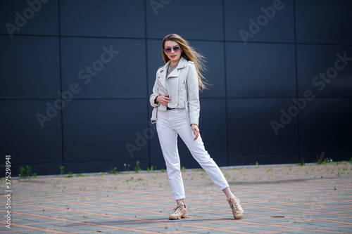 Stylish young woman with long blond hair of European appearance with a smile on her face. Girl in a white jacket and white jeans a warm summer sunny day on a background of a gray building