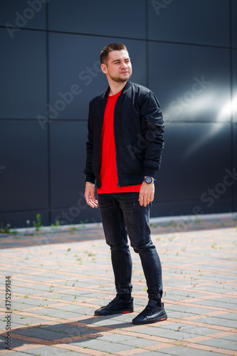 A young man dressed in casual style, a black jacket, jeans, a red T-shirt.  He is against a gray wall Stock Photo | Adobe Stock