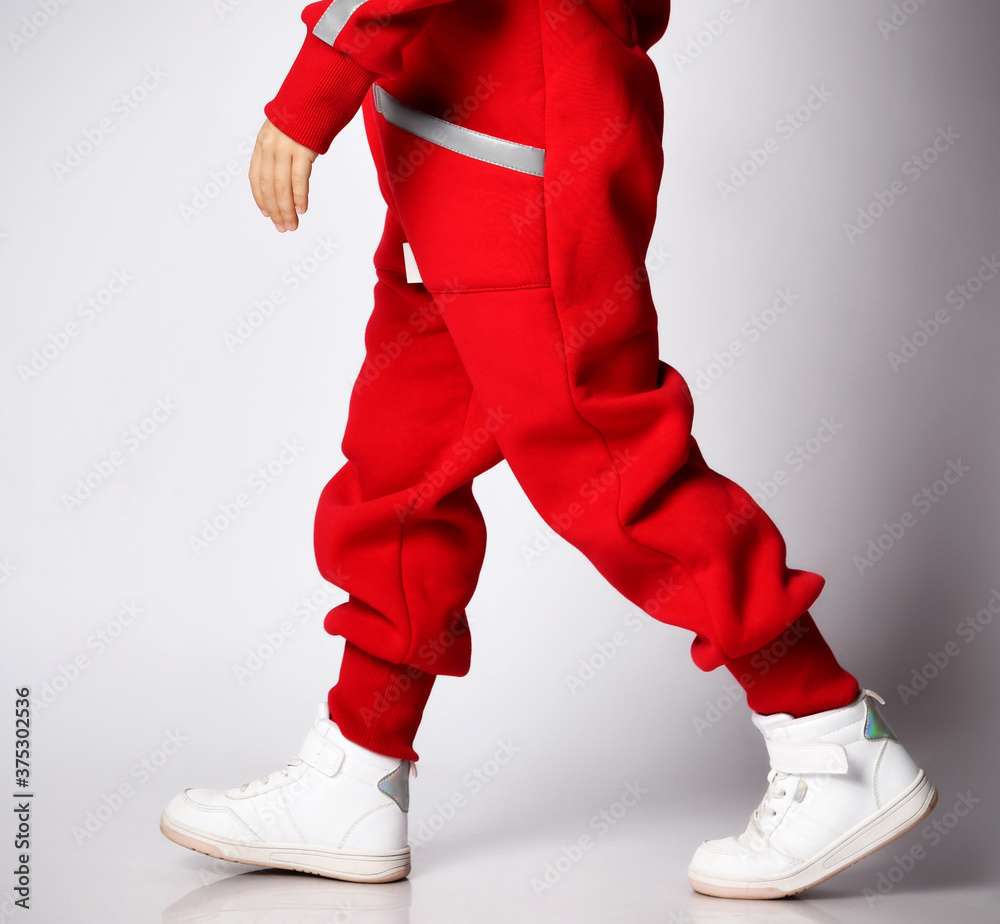 Portrait of cute kid in red overall jumpsuit and white sneakers passing us by, walking