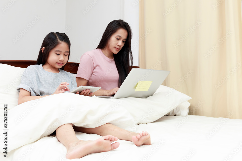 Asian mother and daugther looking laptop and tablet on bed in bedroom. Business women woking with laptop at home with her daugther smiling enjoy with tablet on bed. Work at home concept. Asian mother and daugther looking laptop and tablet on bed in bedroom. Business women woking with laptop at home with her daugther smiling enjoy with tablet on bed. Work at home concept.