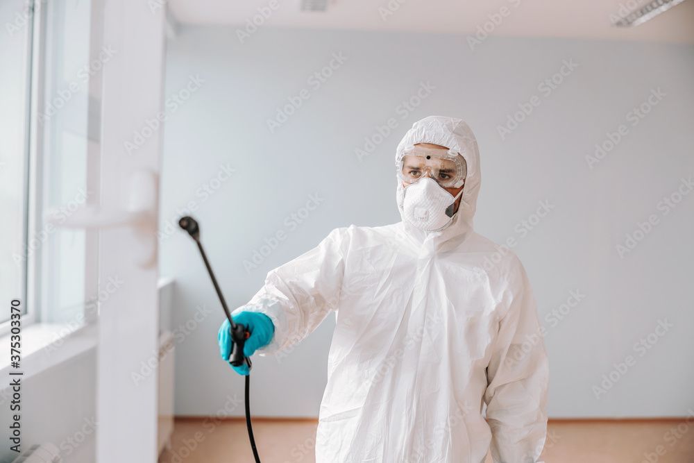 Worker in sterile uniform, with mask and gloves spraying with disinfectant window in school.