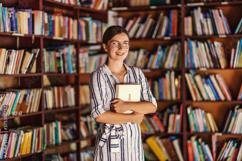 Fotografie Young attractive smart smiling college girl standing in library and holding book while looking at camera