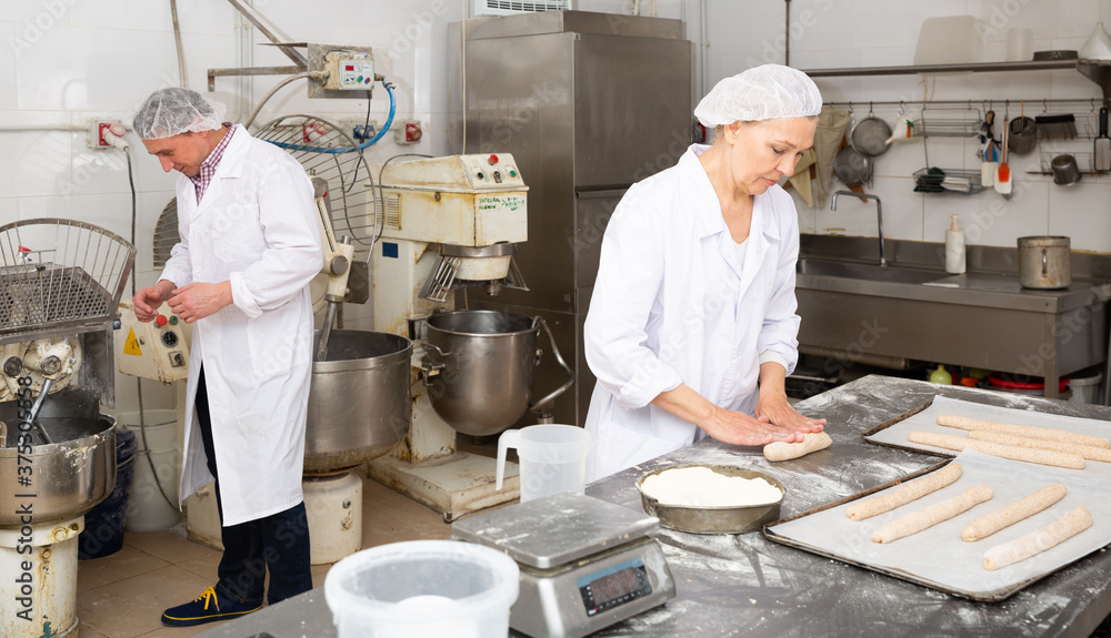 Mature woman professional baker standing at work table and forming dough for baking baguettes