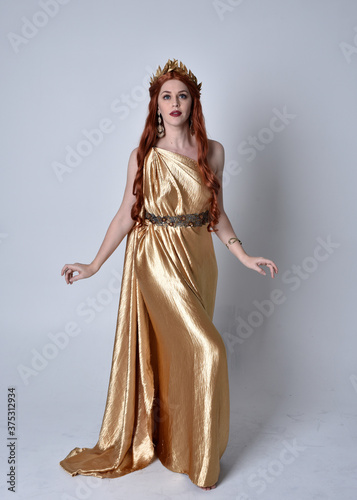 Full length portrait of girl with red hair wearing long grecian toga and golden wreath. Standing pose iisolated against a grey studio background.