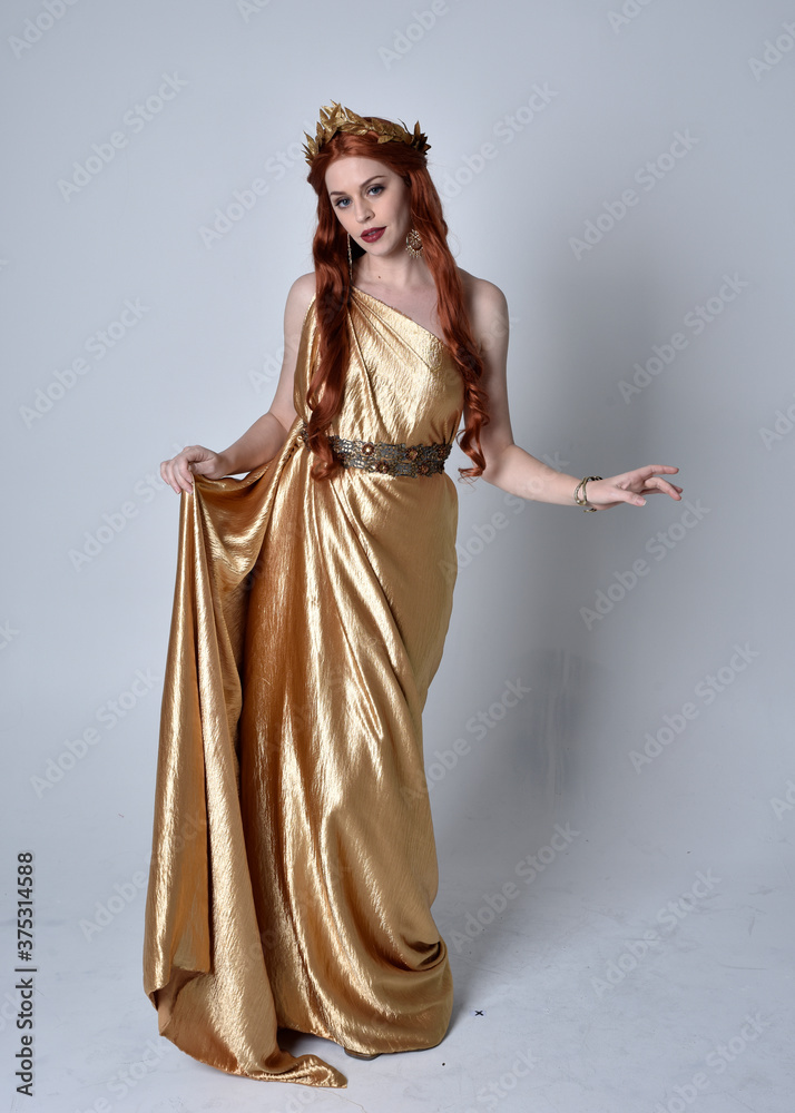 Full length portrait of girl with red hair wearing long grecian toga and golden wreath. Standing pose iisolated against a grey studio background. Full length portrait of girl with red hair wearing long grecian toga and golden wreath. Standing pose iisolated against a grey studio background.