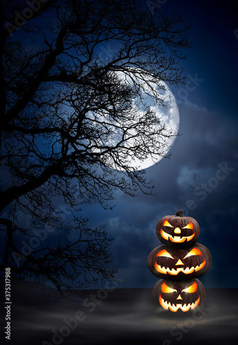 A pile of three spooky halloween pumpkins, Jack O Lantern, with evil face and eyes under the silhouette of a tree at night with a full moon and misty sky. photo