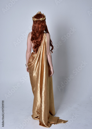 Full length portrait of girl with red hair wearing long grecian toga and golden wreath. Standing pose with back to the camera,  isolated against a grey studio background. photo