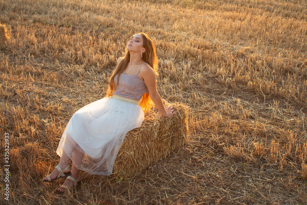 Happy woman sitting on hay stack walking in summer evening, beautiful romantic girl with long hair outdoors in field at sunset Happy woman sitting on hay stack walking in summer evening, beautiful romantic girl with long hair outdoors in field at sunset