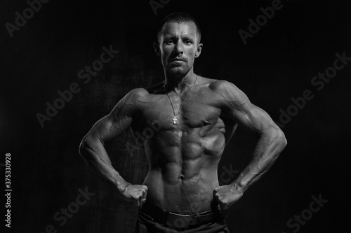 Black and white portrait of a slender athletic man with a naked muscular torso against a dark background.