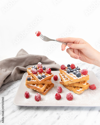 Golden Belgian waffles with raspberries and blueberries topped with powdered sugar. Woman hand holding a fork. Grey plate on marble table. Bakery or cafè breakfast. Food styling, front view close-up