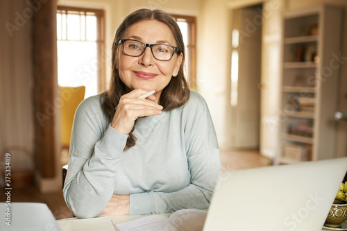 Portrait of gray haired mature businesswoman in eyewear using portable computer for remote work, studying documentation. Attractive retired female teacher holding pen, checking home assignment