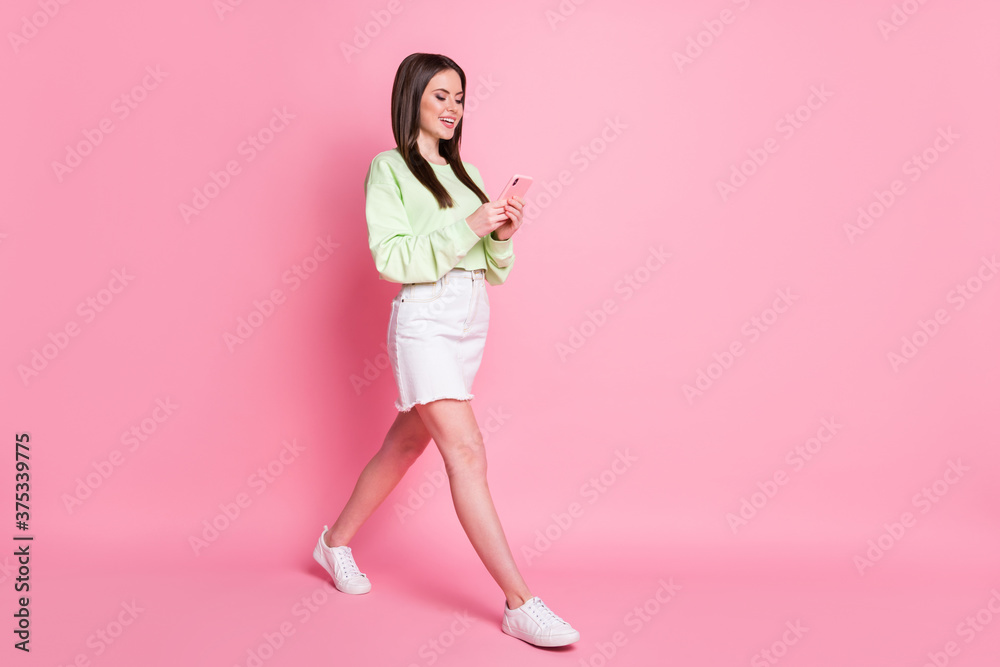 Full size profile side photo of positive girl blogger walk go use smartphone read social media news wear green white trendy jeans sweater sneakers isolated pastel color background Full size profile side photo of positive girl blogger walk go use smartphone read social media news wear green white trendy jeans sweater sneakers isolated pastel color background