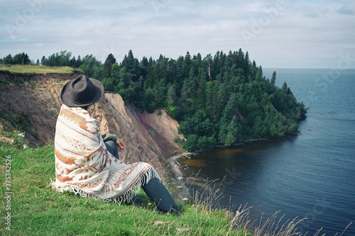 Lonely woman in felt hat and woolen poncho sitting on grassy seashore and looking at wooded cape. Travel, adventure, feeling nature, folk ethnic lifestyle. Lake Onega, Russian North.