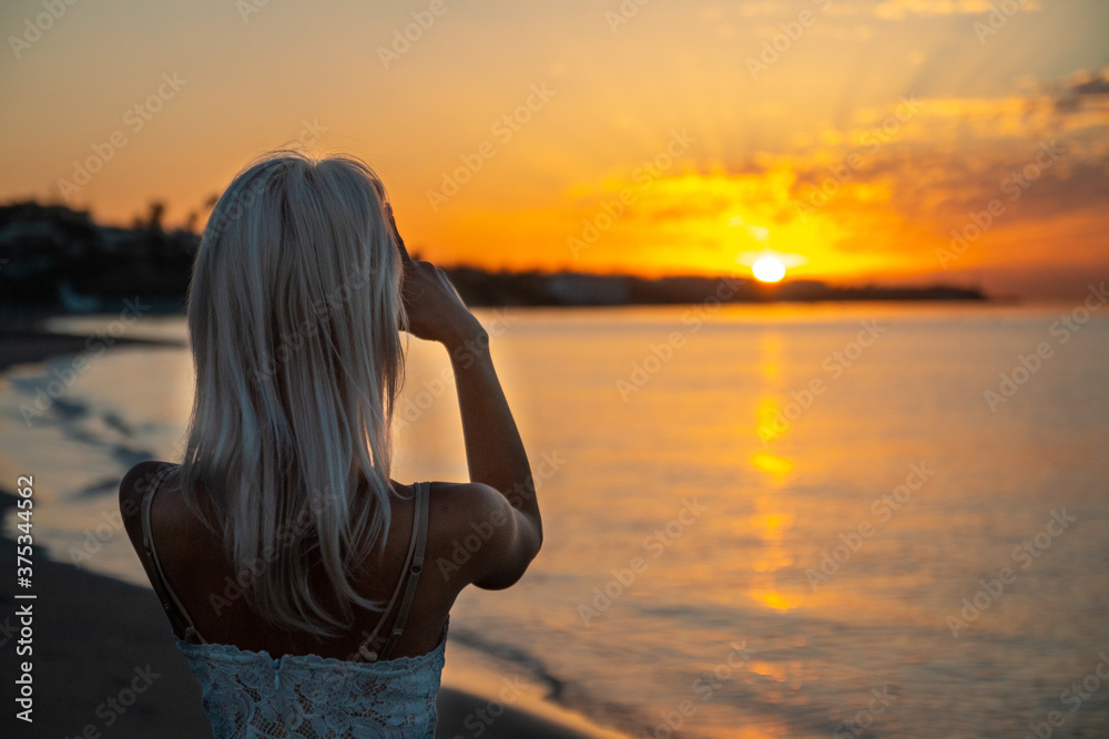 Young blonde women taking a photo of a sunrise on the beach over the sea Young blonde women taking a photo of a sunrise on the beach over the sea