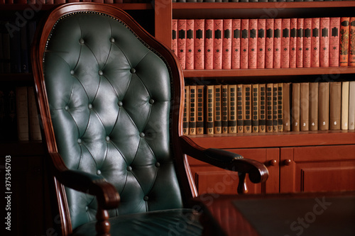 Dark solid interior of old library with books on wooden shelves. Vintage classic chair green leather and red wood, antique table, luxury design with expensive natural details. Indoor, low exposure.