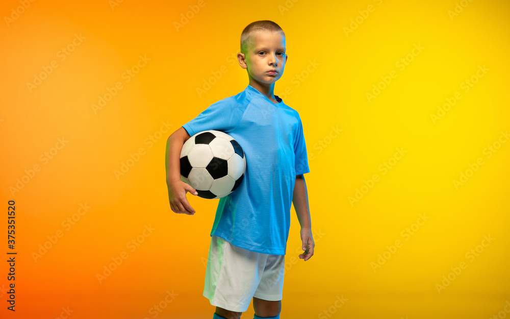 Confidence. Young boy as a soccer or football player in sportwear practicing on gradient yellow studio background in neon light. Fit playing boy in action, movement, motion at game. Copyspace. Confidence. Young boy as a soccer or football player in sportwear practicing on gradient yellow studio background in neon light. Fit playing boy in action, movement, motion at game. Copyspace.